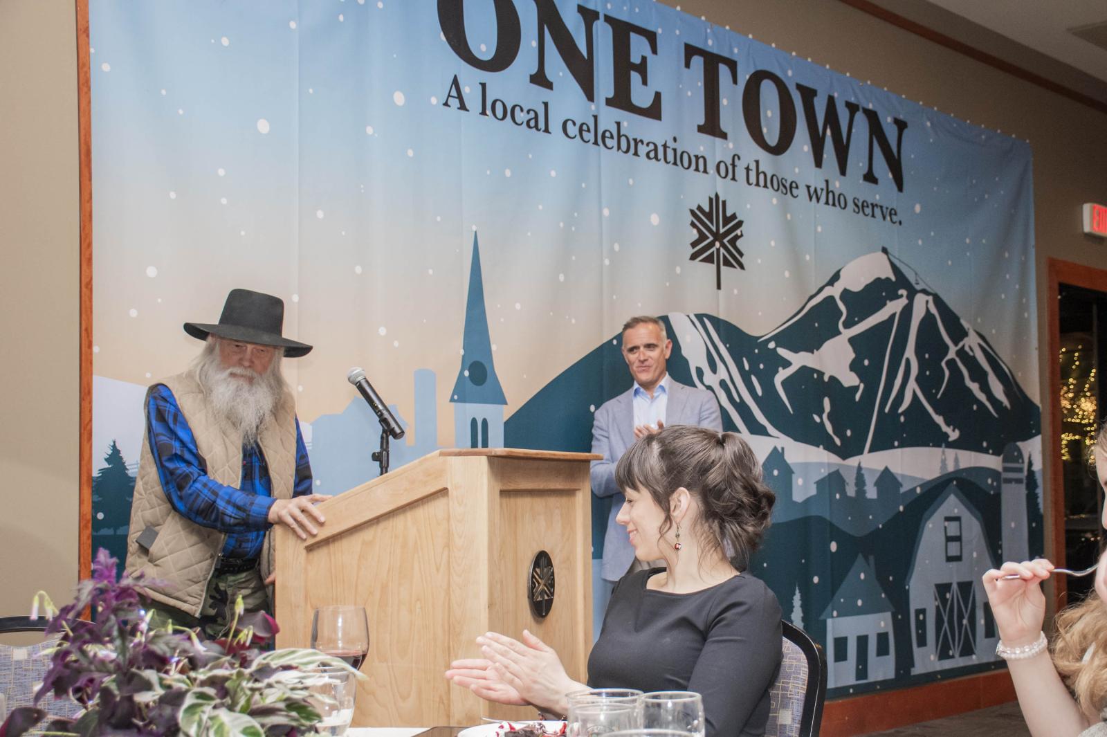 Man with long beard at podium, banner with mountains and text "ONE TOWN" in background.