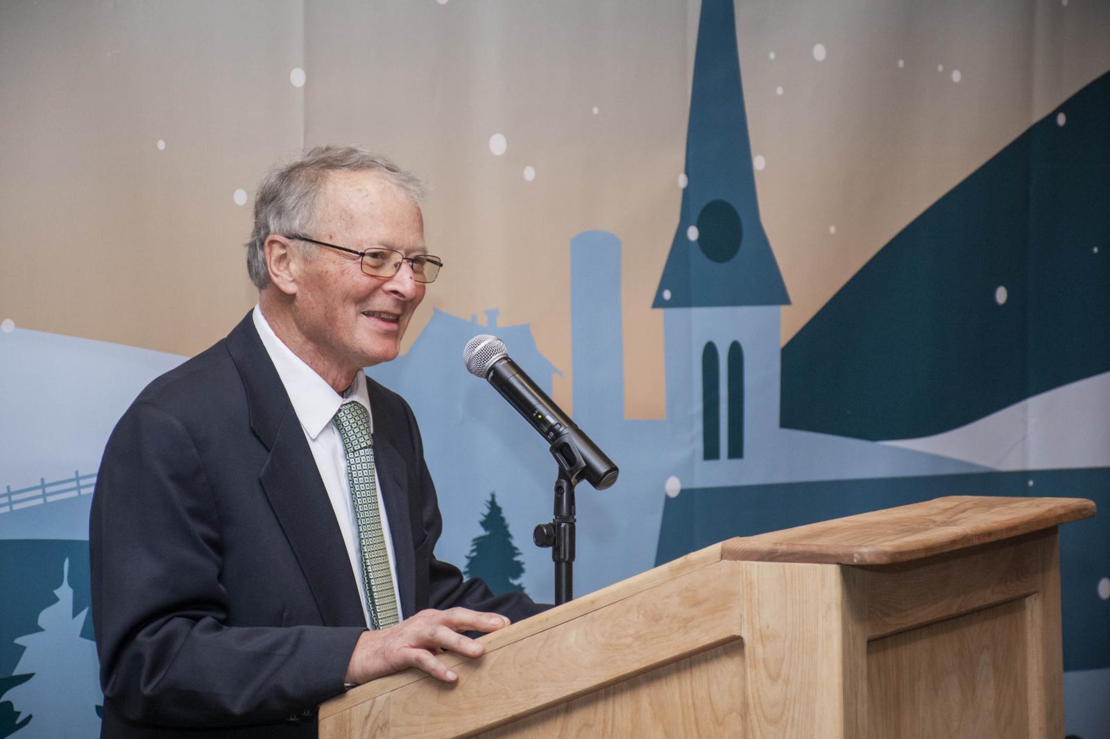 Elderly man speaking at a podium, church silhouette in the background.