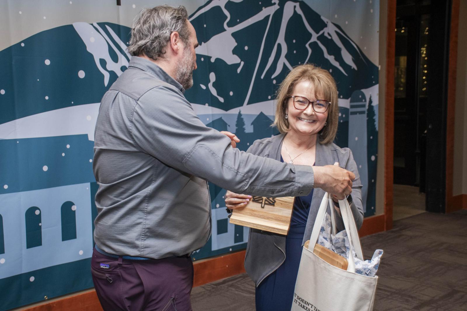 Man and woman exchange items, smiling in a room with a mountain backdrop.