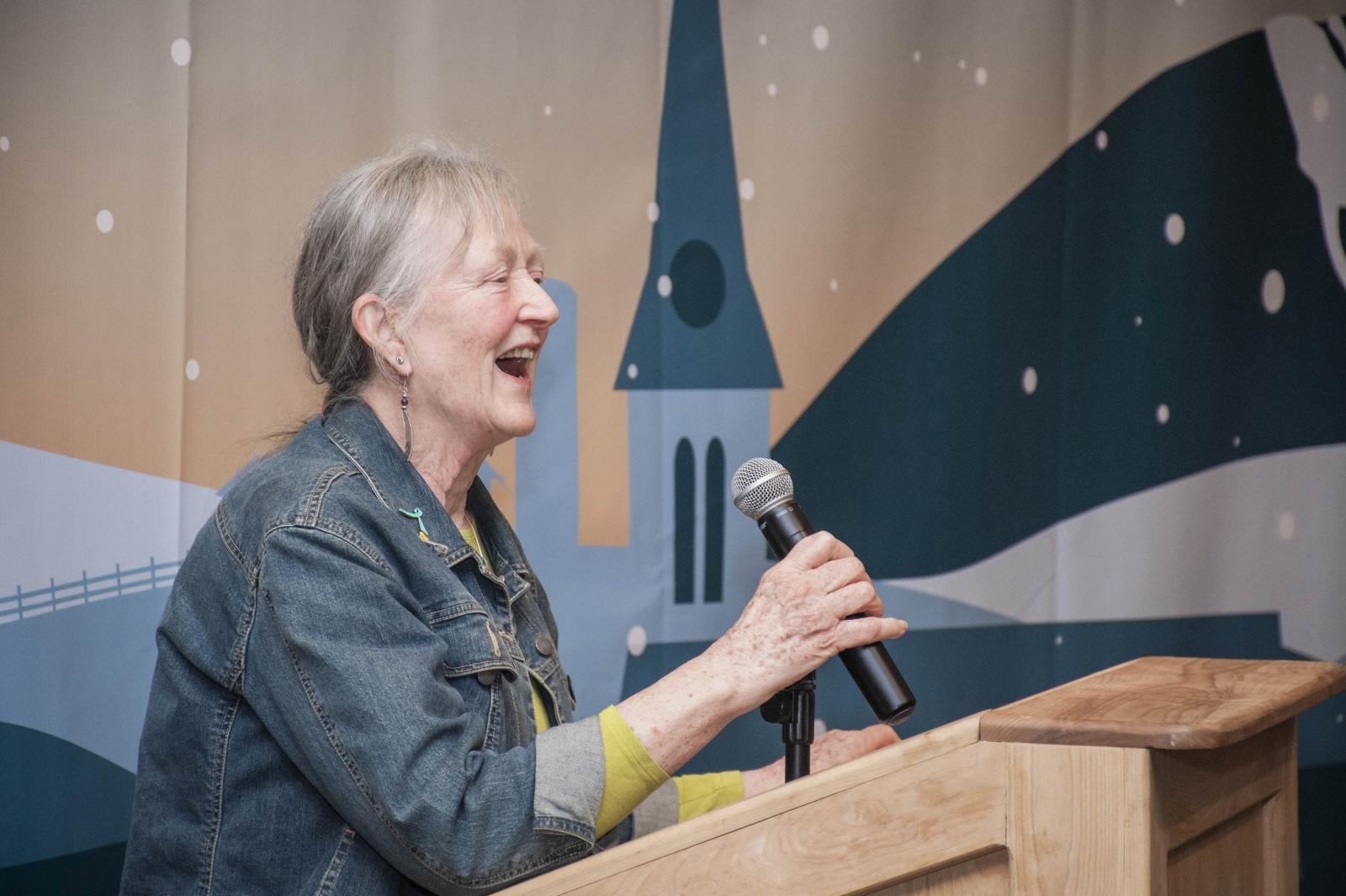 Elderly woman laughing while speaking into a microphone at a podium.