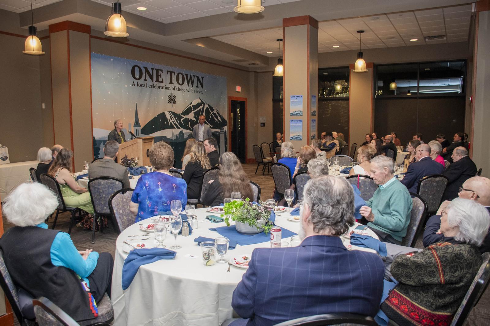 Audience seated at round tables, listening to a speaker in a conference room.