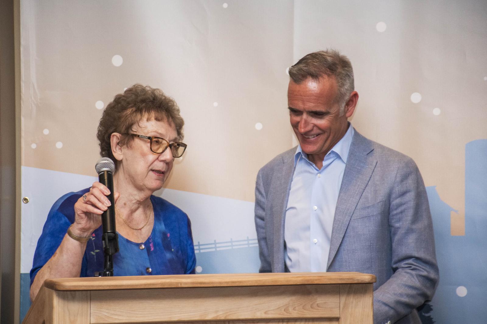Elderly woman speaks at podium, man in suit smiles beside her.