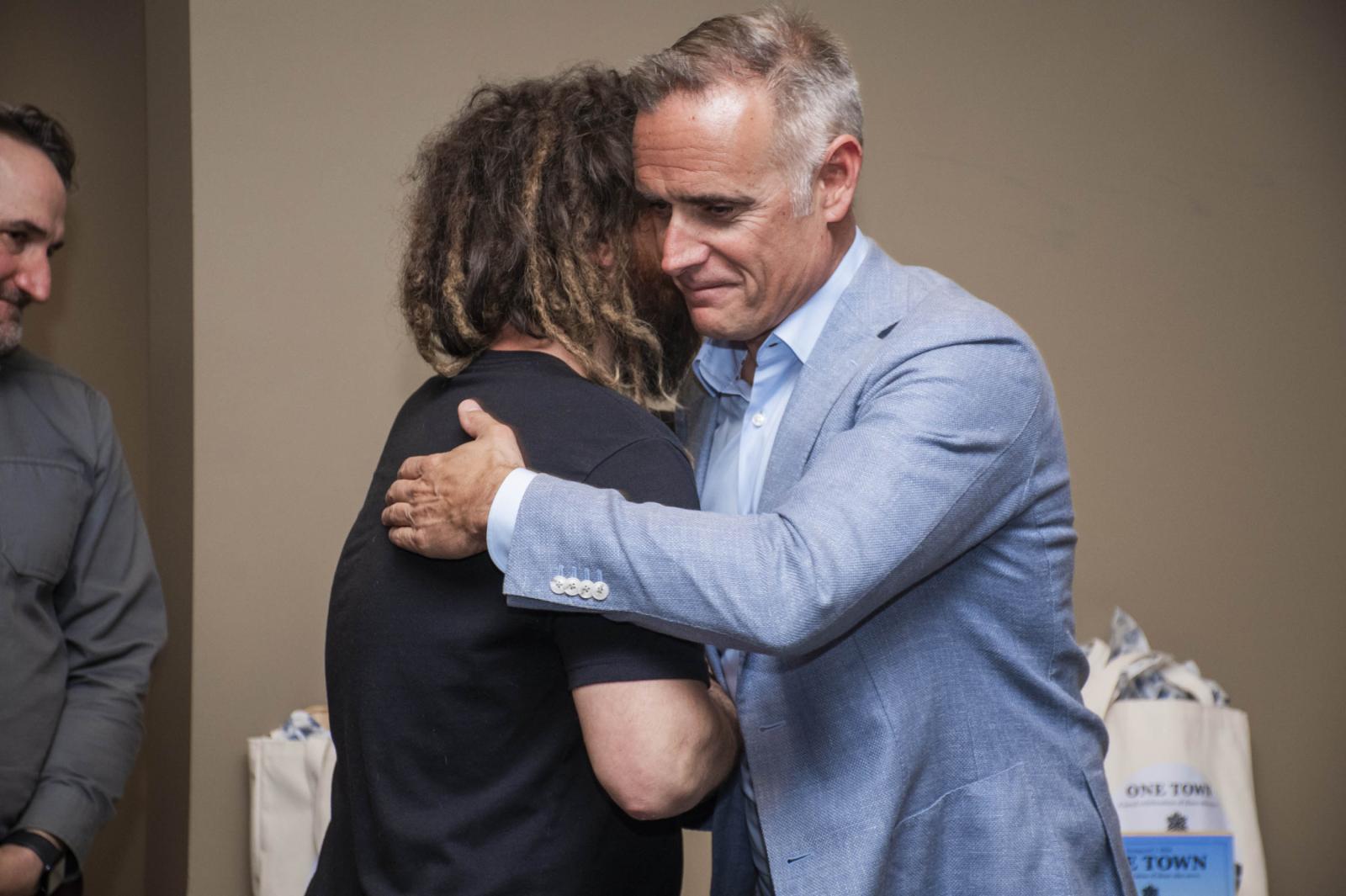 Man in a blue suit hugs another man with long hair in a black shirt.