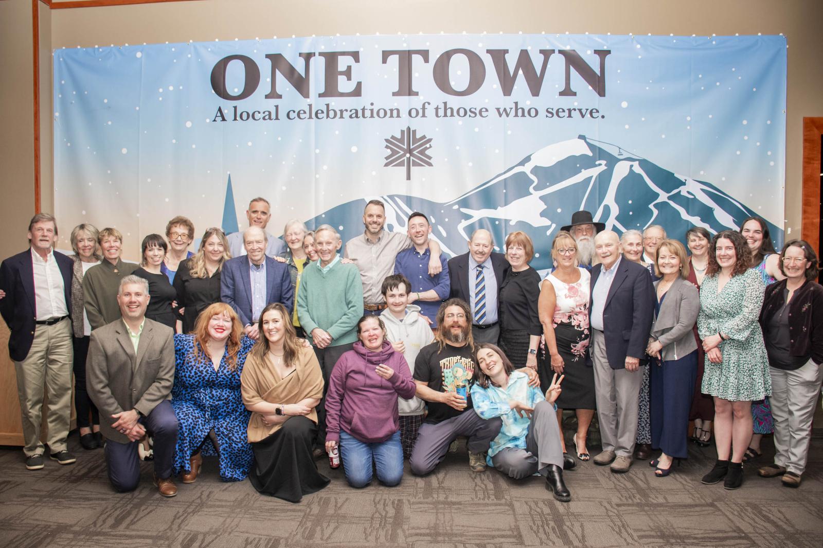 Group of people posing in front of a "ONE TOWN" banner with a mountain graphic.