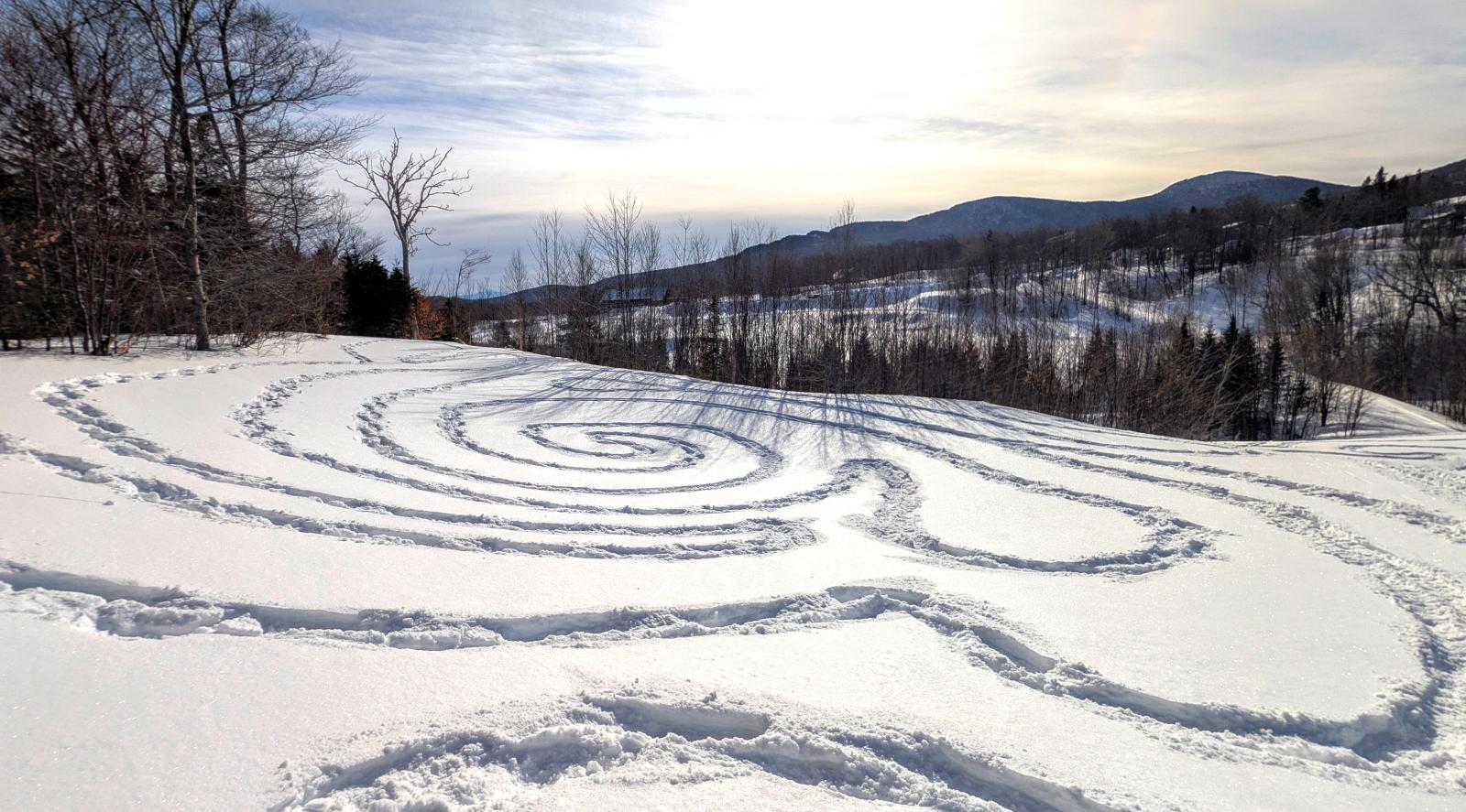 Snow-covered field with spiral patterns, hills, and a blue sky in the background.
