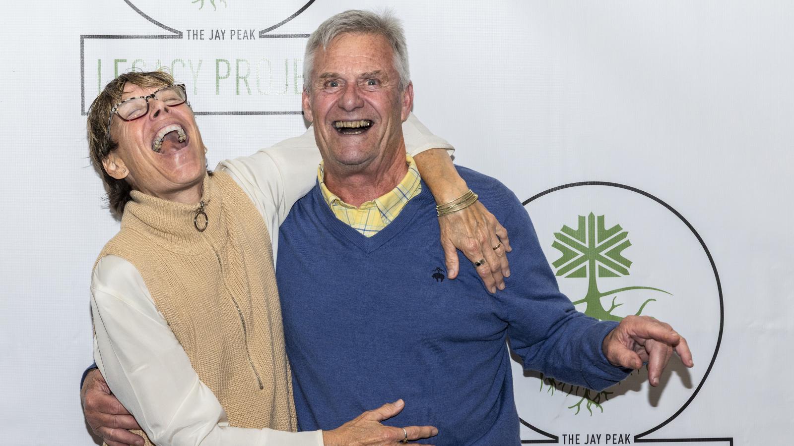 Elderly couple laughing joyfully, arm in arm, against a branded backdrop.