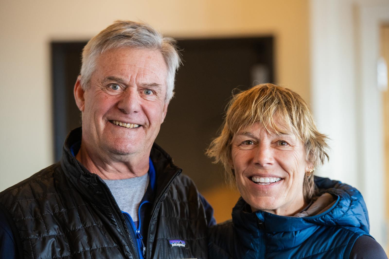 Older couple smiling indoors, wearing jackets.
