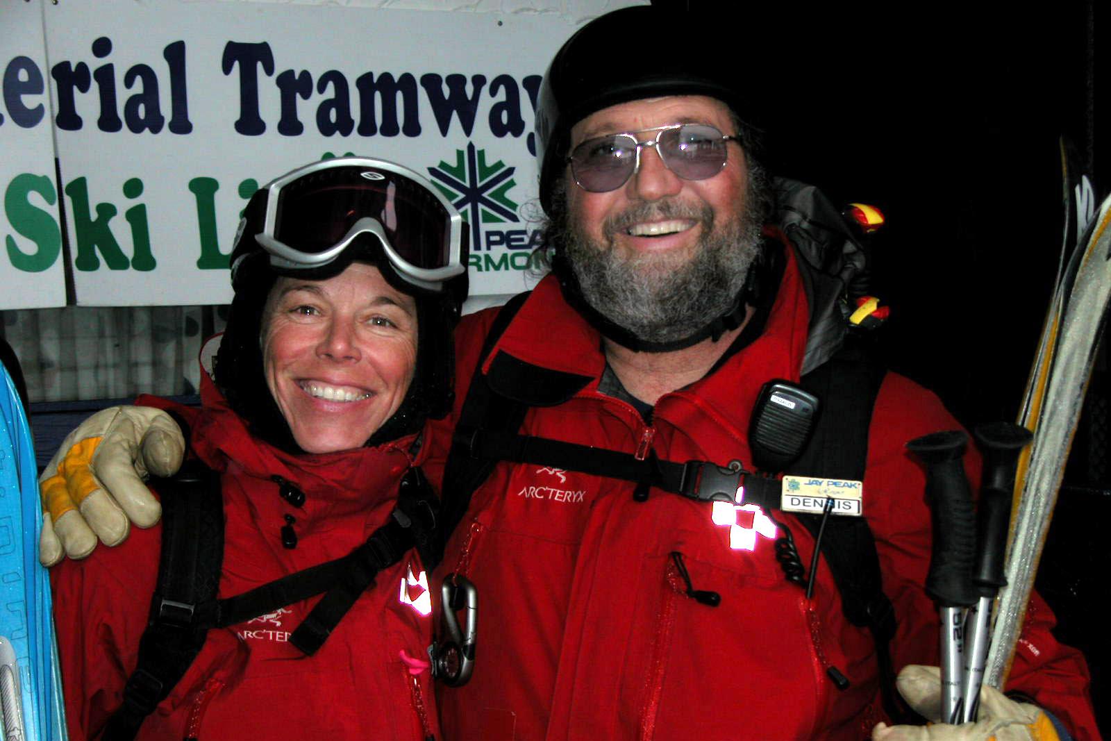 Two smiling skiers in red jackets, one holding skis, standing together at night.