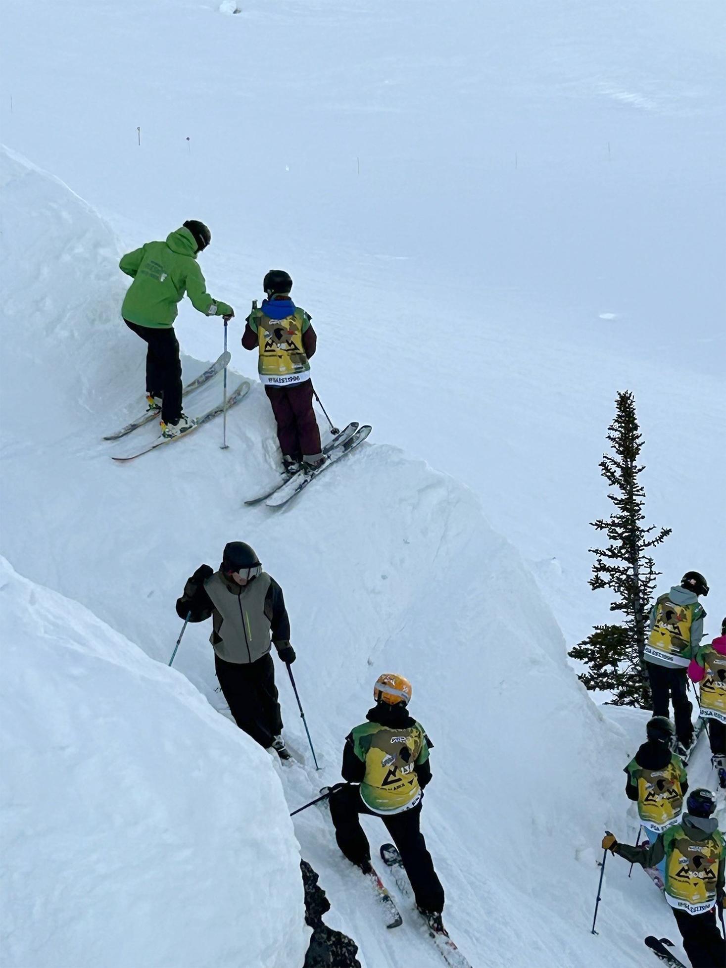 Skiers on a snowy slope, some in green jackets, near a small tree.