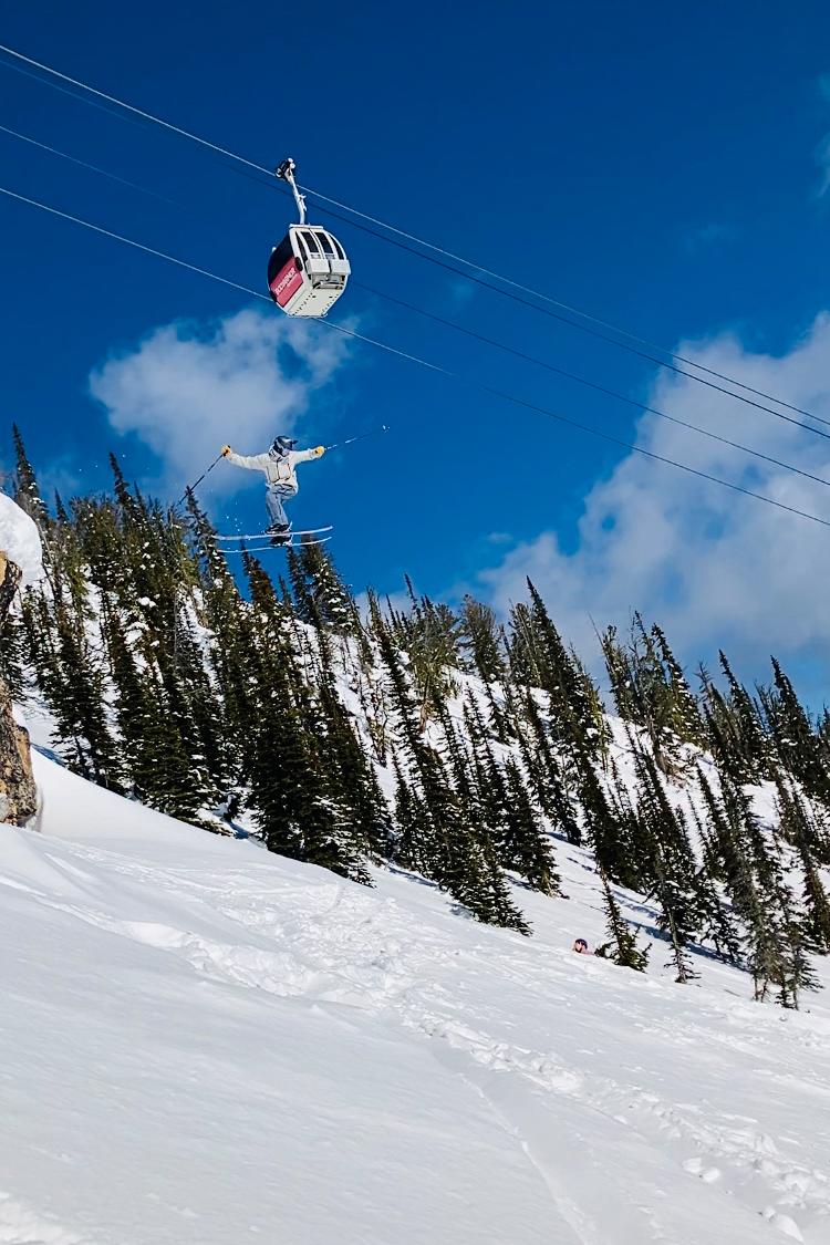 Snowy slope with a gondola in the sky, trees, and skiers below.