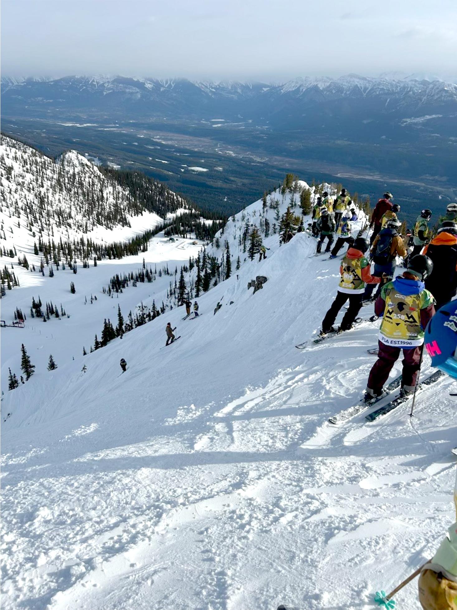 Skiers on a snowy mountain slope overlooking a vast, distant valley.