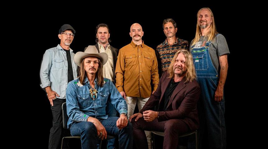 Seven men pose, one seated, in varied attire against a black background.