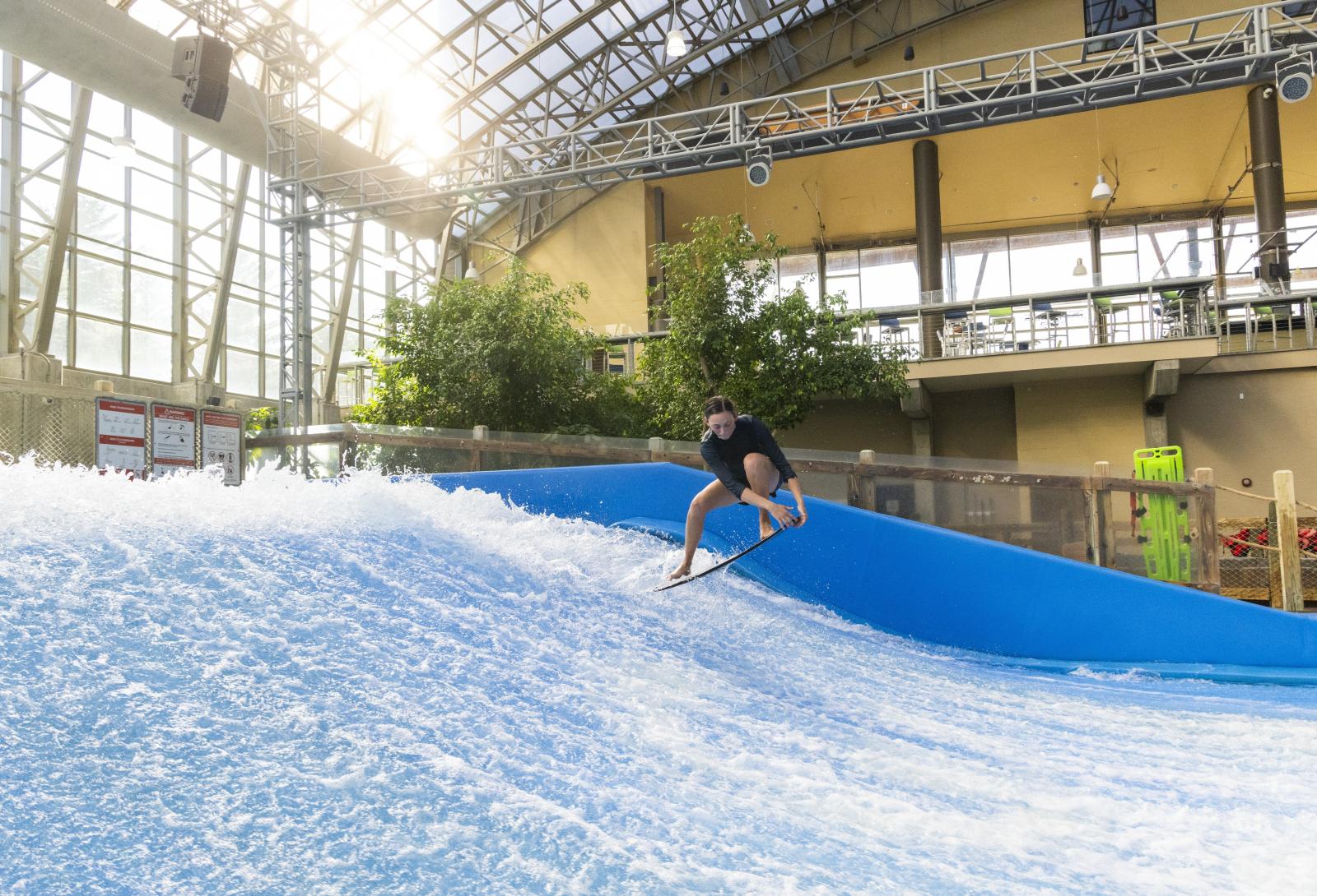 Indoor surfing on artificial wave, surfer balances under glass roof.