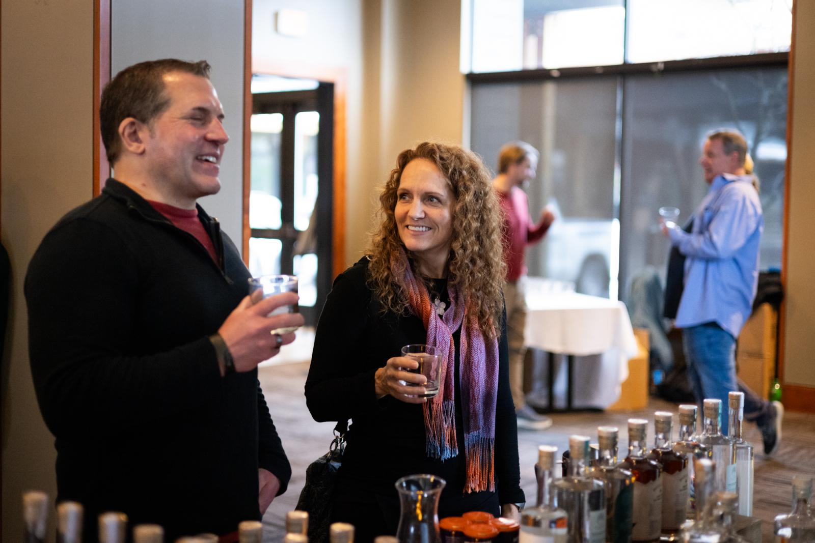 Two people smiling and tasting beverages at a bottle display table.