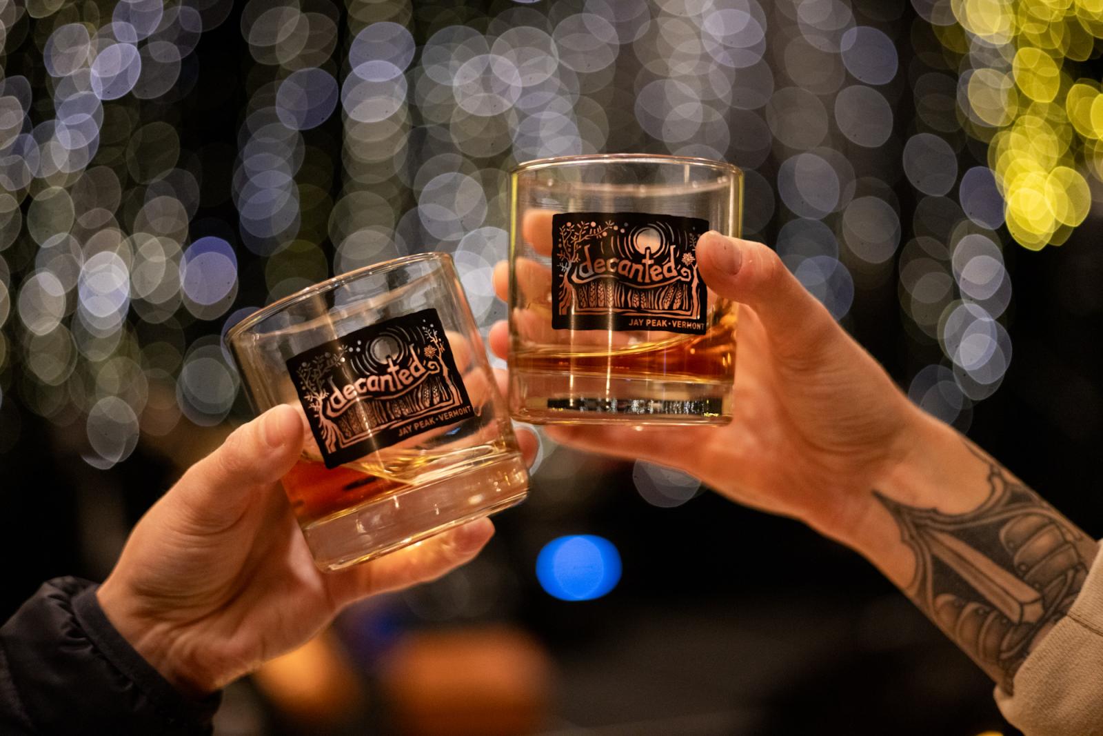 Two hands toasting with glasses of whiskey against a blurred light backdrop.