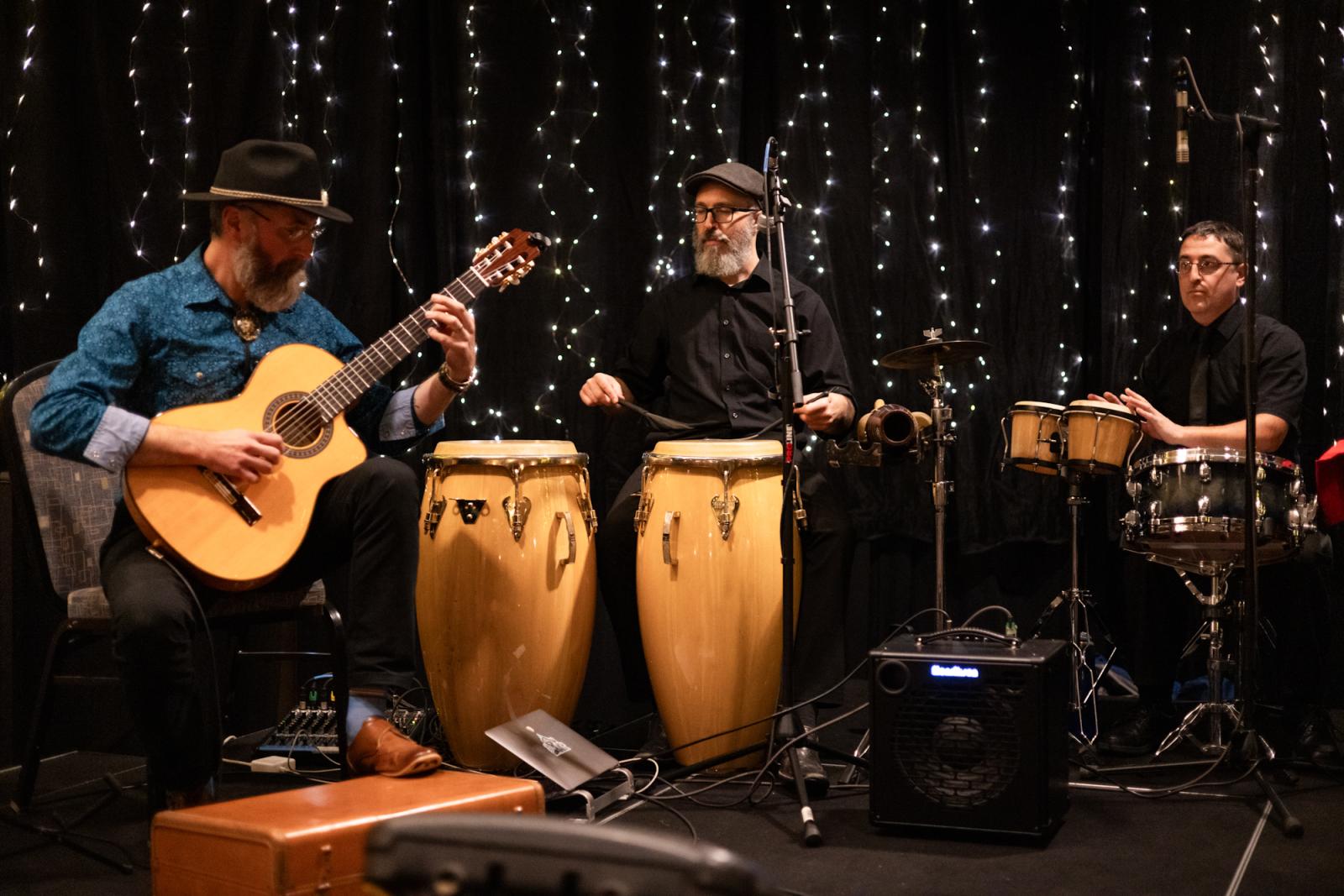 Musicians performing with guitar, congas, and drums in dimly lit setting.
