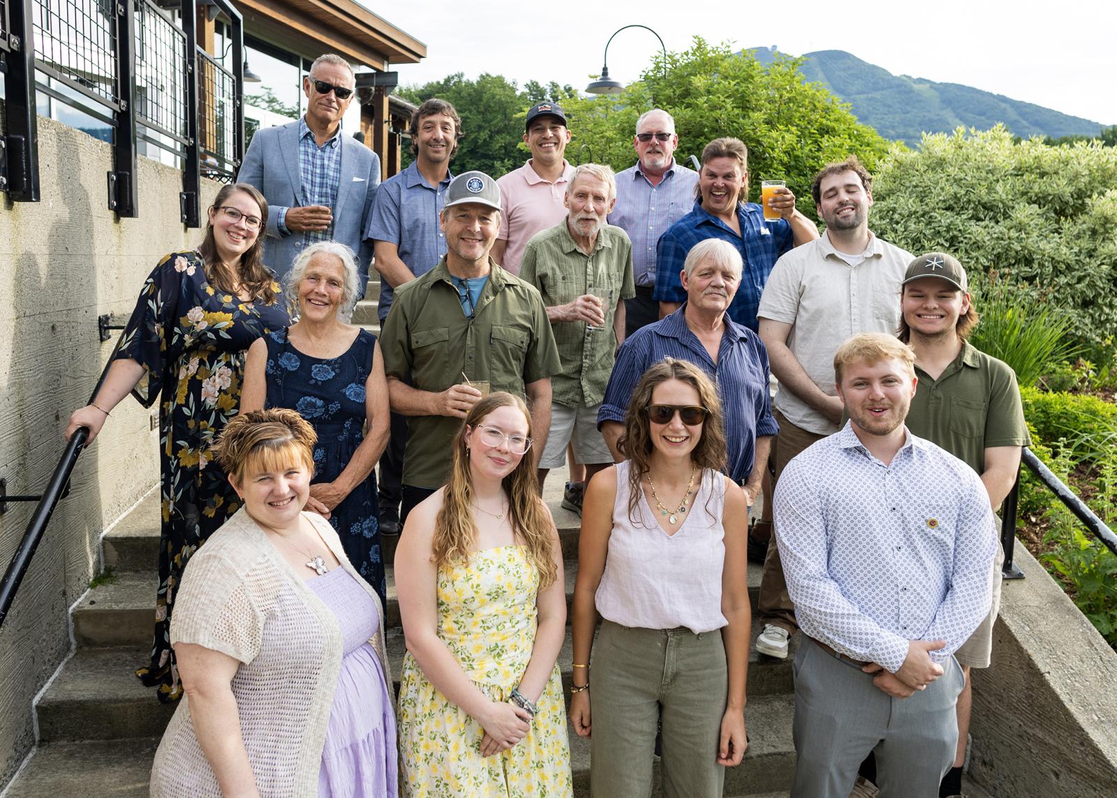 Group of people posing outdoors on steps, smiling, with greenery and mountains in the background.