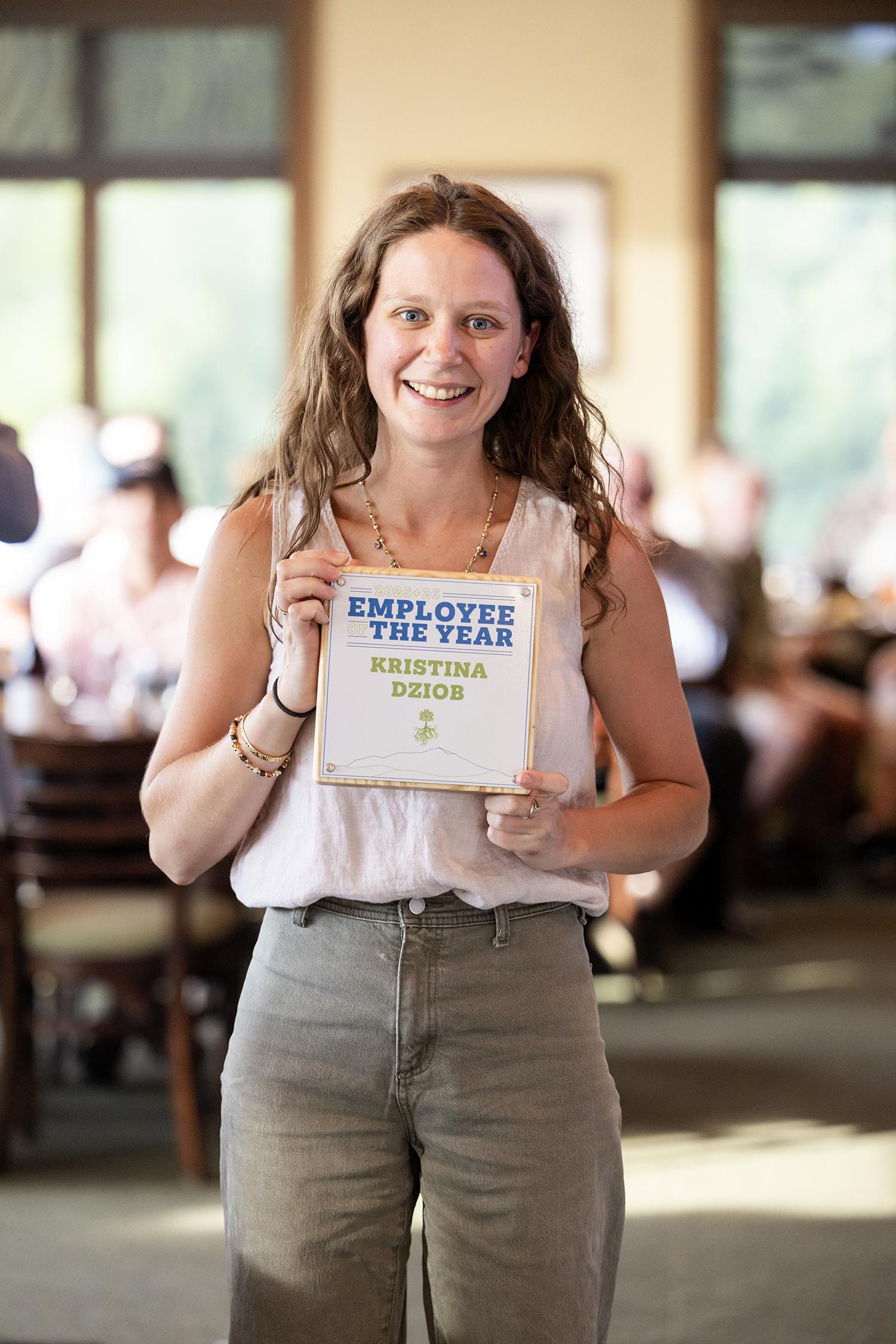 Woman smiling, holding a framed award in a bright room.