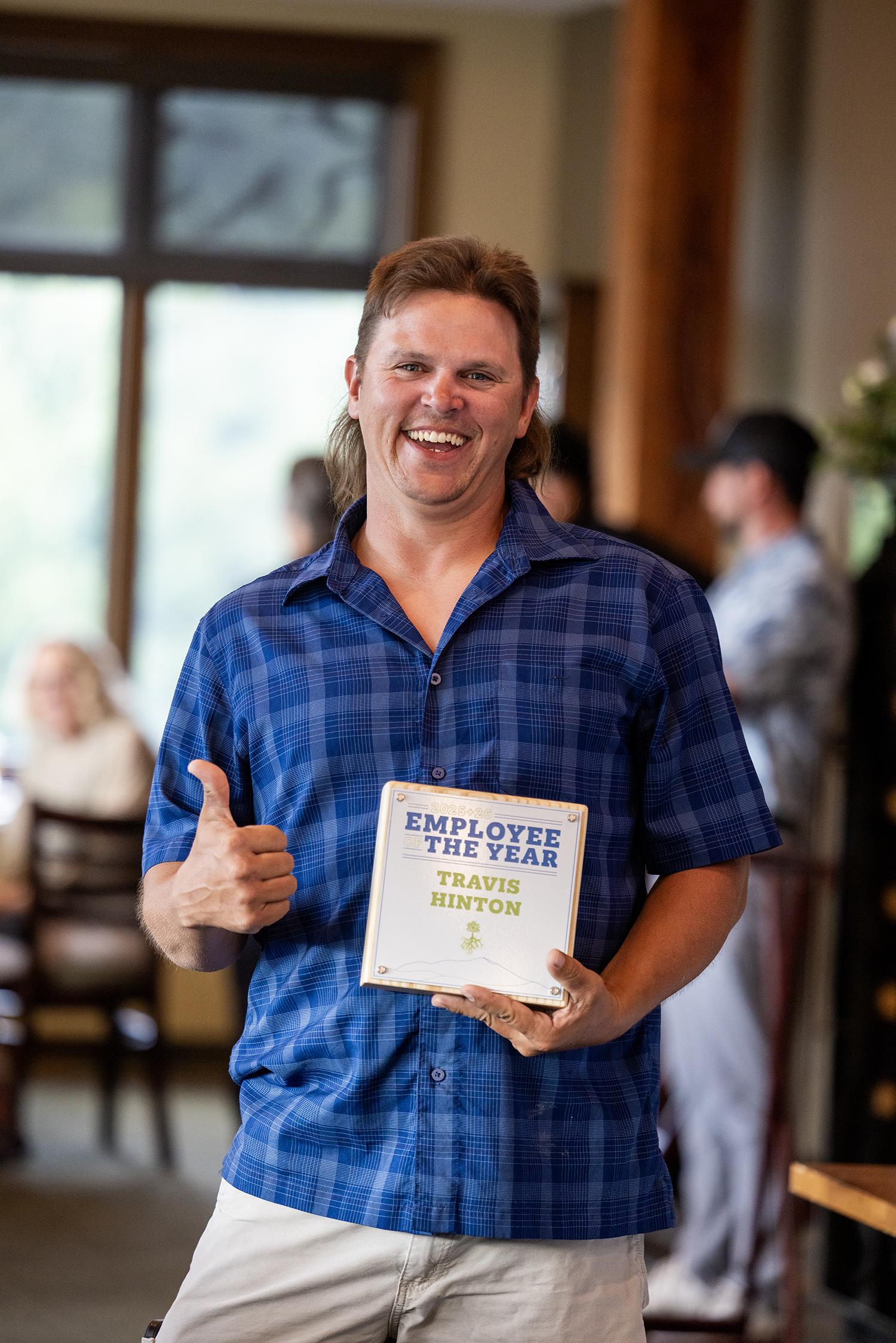 Man smiling with thumbs up, holding a sign, wearing a blue plaid shirt indoors.