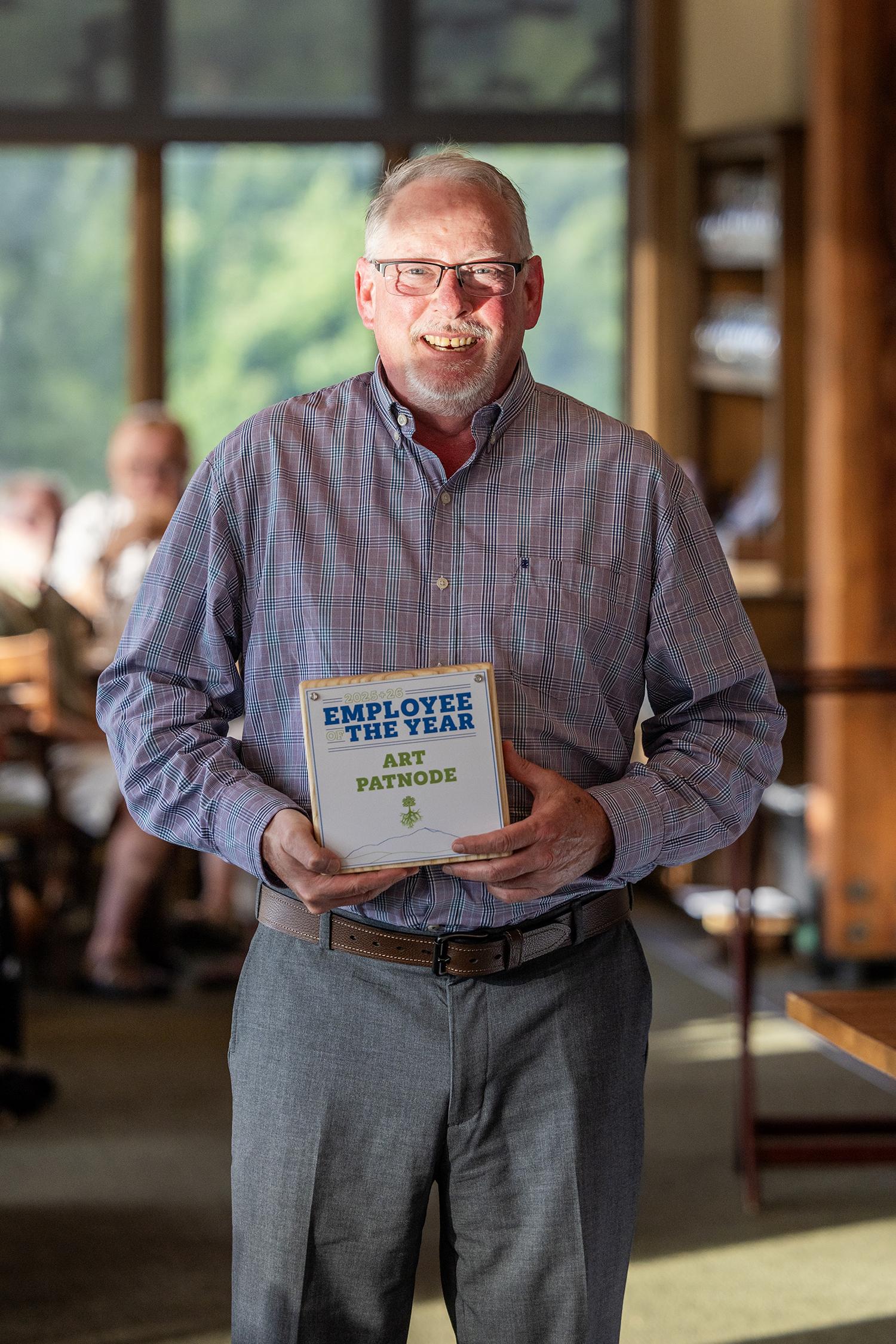 Man in glasses holding an award in a sunlit room.