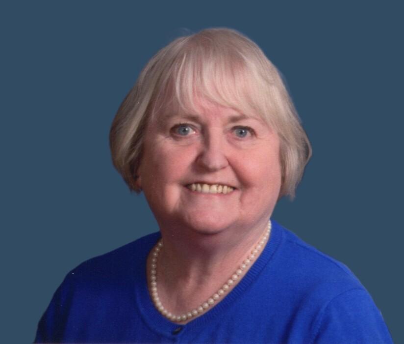 Smiling woman with short white hair, wearing a blue top and pearl necklace.