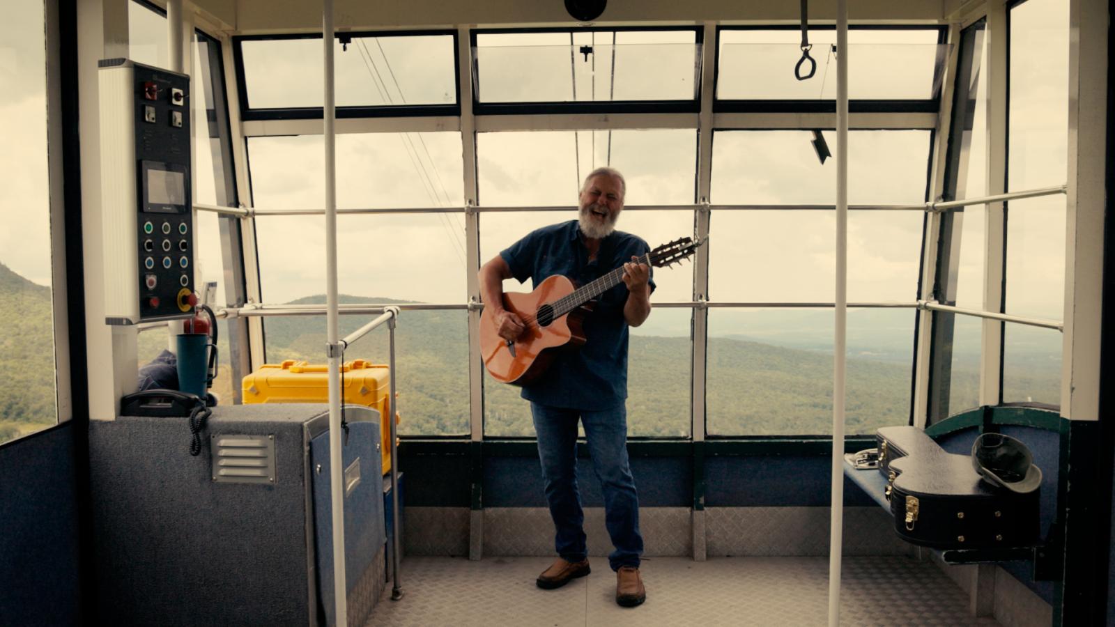 Musician playing guitar inside a cable car with mountain views.