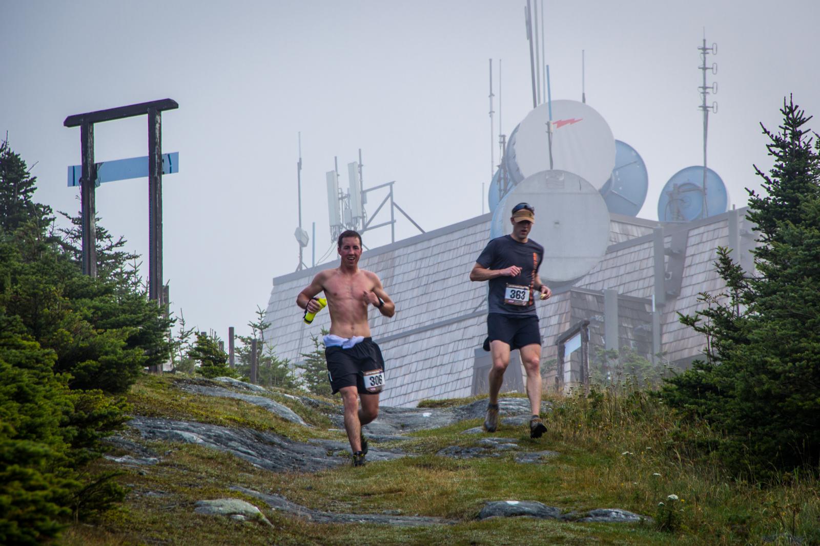 Two men running on a rocky trail beside communication towers.