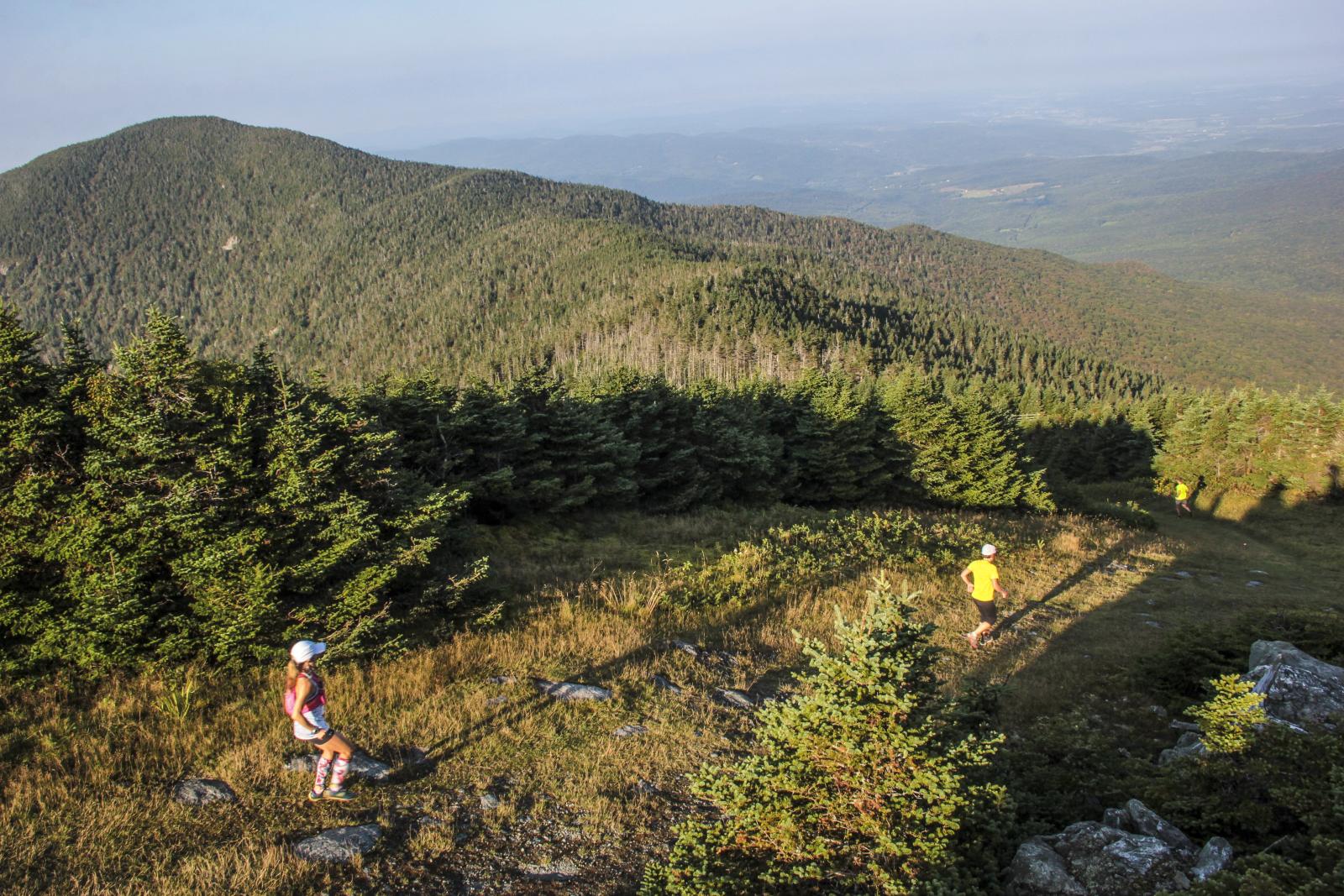 Hikers on a rocky mountain trail overlooking forested hills at sunset.