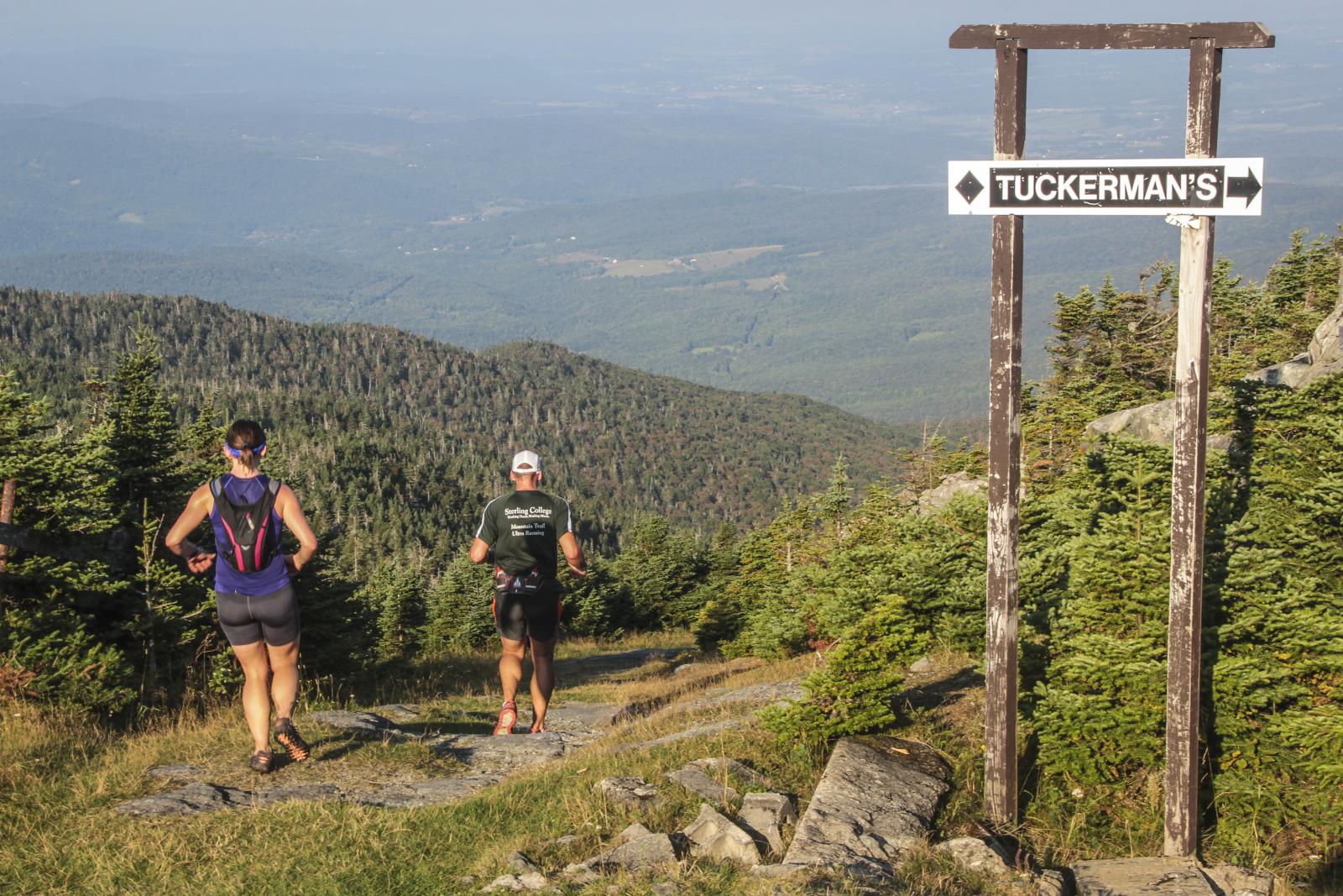 Two hikers on a mountain trail beneath a wooden signpost.