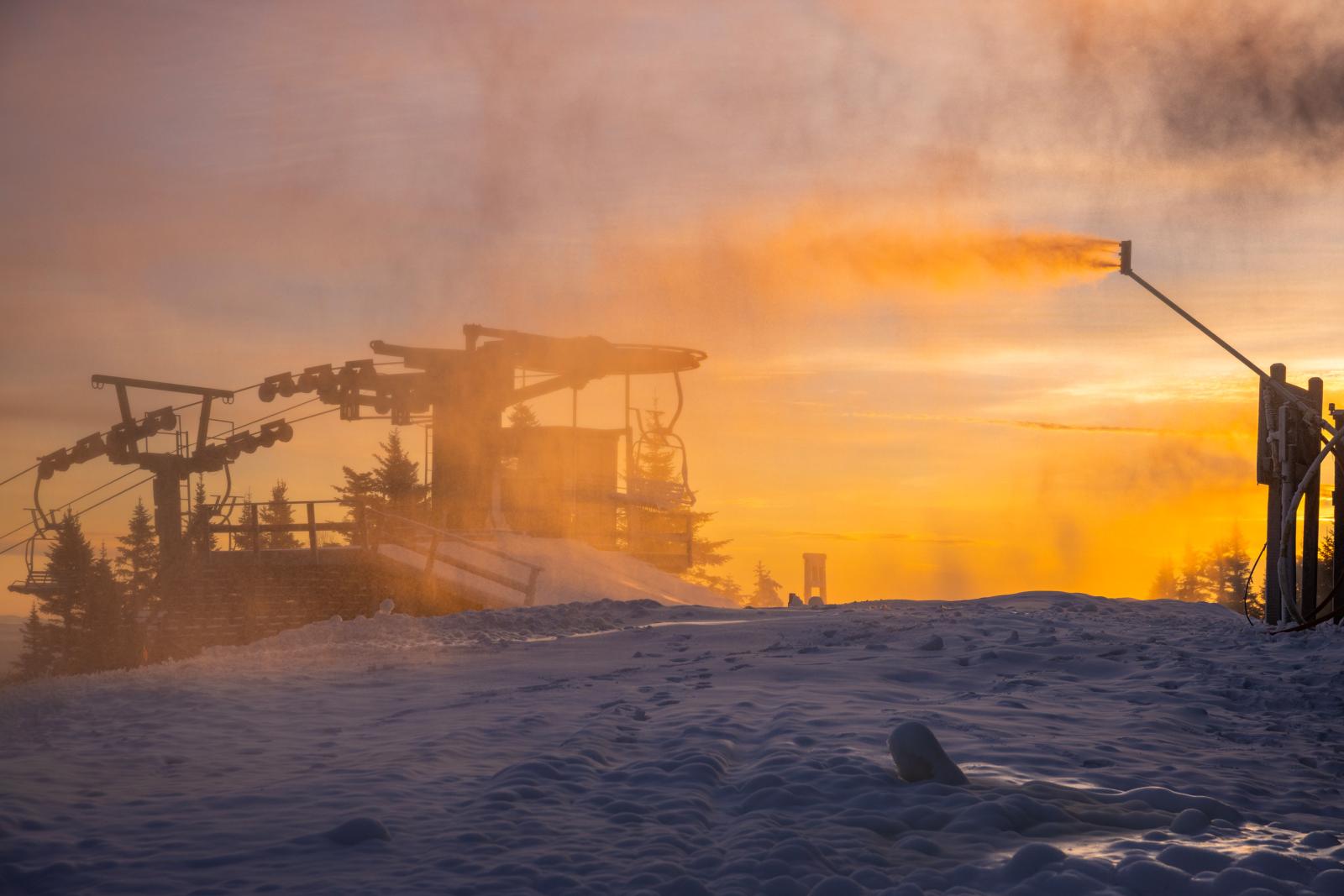 Snow-covered ski lift and and snowmaking silhouetted against a golden sunrise.