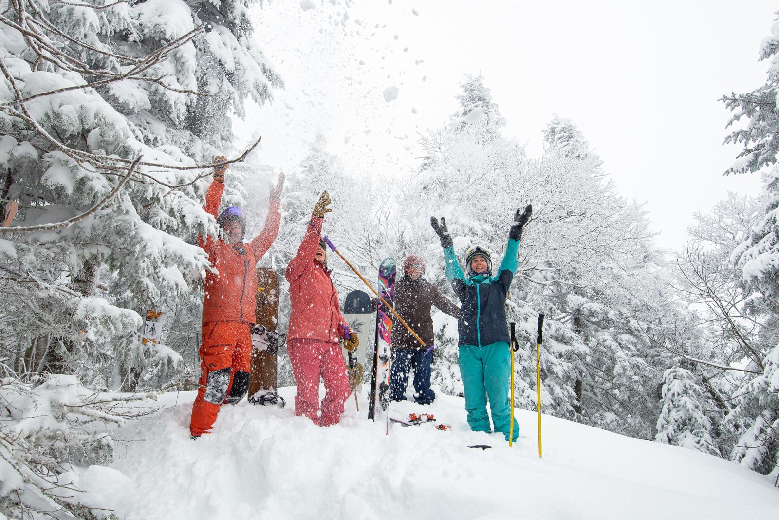 Four friends in colorful snowsuits celebrate atop a snowy hill.